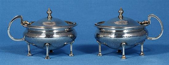 A pair of George V Arts & Crafts silver mustard pots with matching spoons, by Albert Edward Jones, width 90mm, weight 5.1oz/160grms.